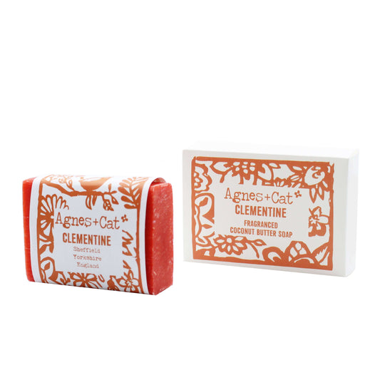 A bar of clementine-scented coconut butter soap next to its packaging box, which has a floral pattern and reads 'Agnes+Cat Coconut Butter Soap - Clementine'.