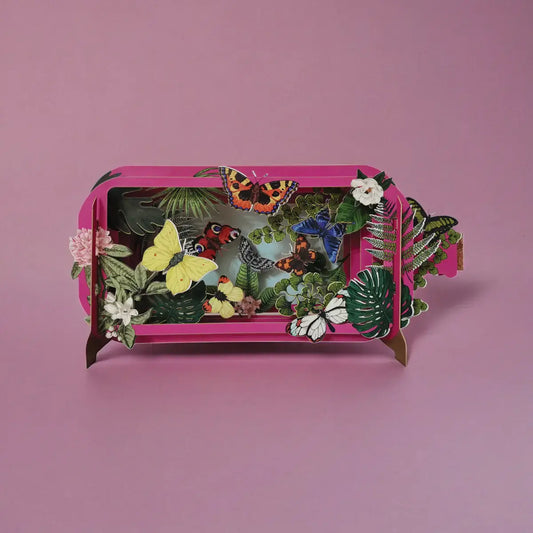 A colorful pop-up greeting card with a butterfly design, featuring a variety of flowers and leaves, set against a pink background.