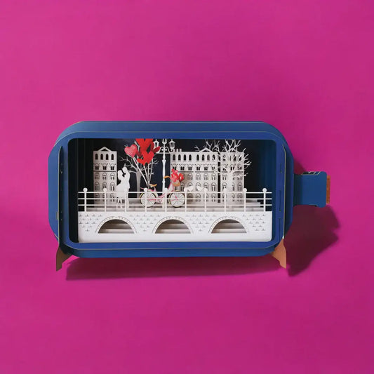 Pop up card with white cut out layers of a town scene and bridge. Cut out image of a couple embracing on the bridge. A bicycle beside them has red flowers in basket and red heart balloons tied to it