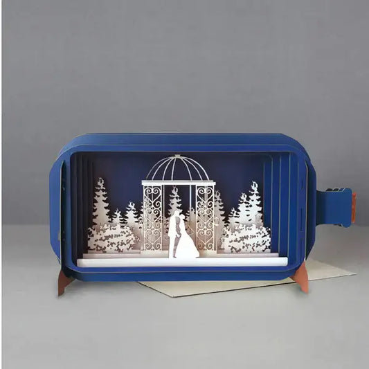 A blue pop-up greeting card displayed open with a delicate design depicting a wedding scene in white layers, including a bride and groom, trees, and a decorative arbour.