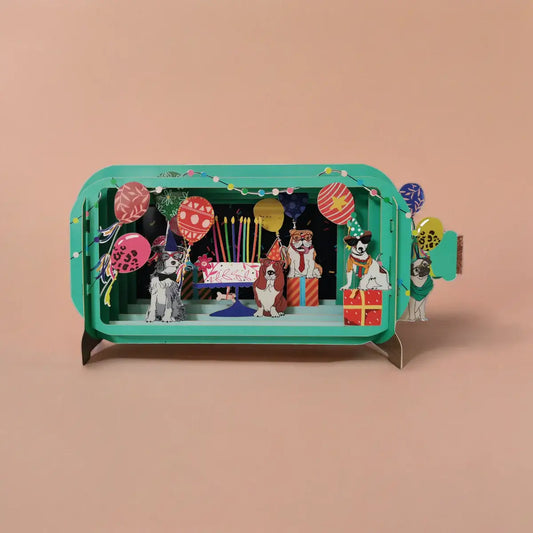 A colorful pop-up greeting card featuring an illustration of dogs at a birthday party with balloons and a cake, housed in a green rectangular box.
