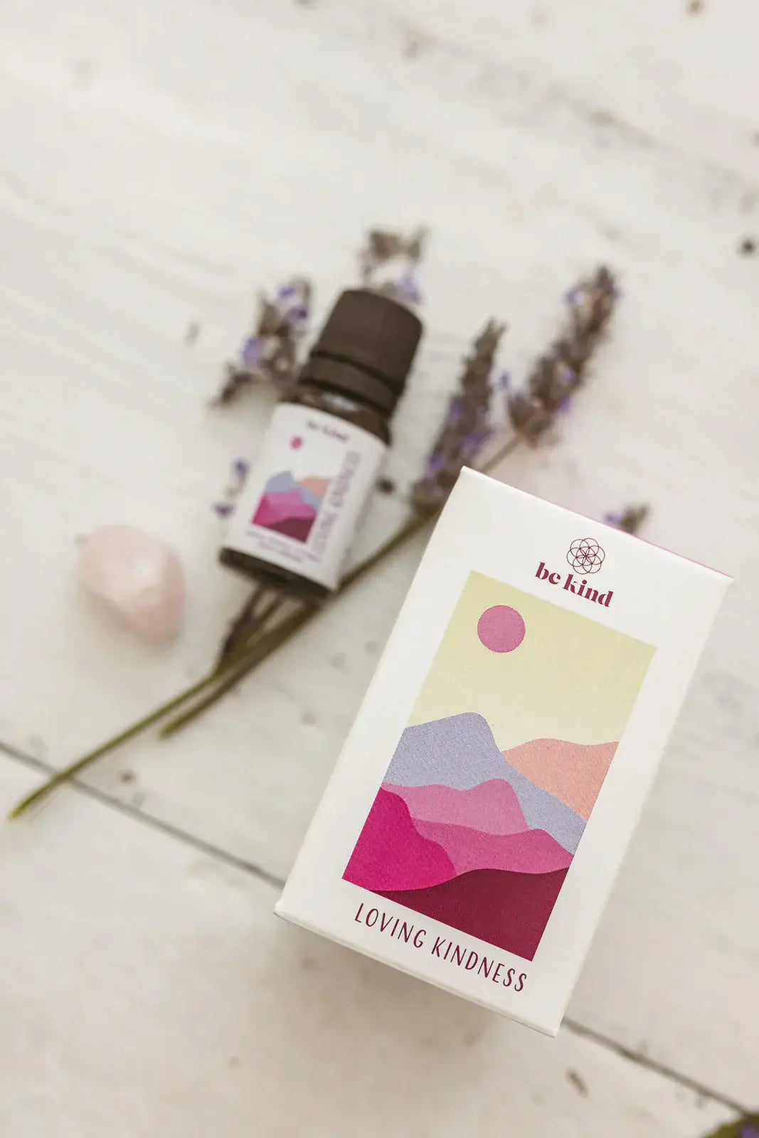 Essential oil bottle and box with 'be kind' and 'Loving Kindness' text on a light wooden surface with lavender flowers.
