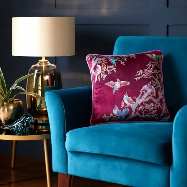 A square magenta velvet throw pillow with a colorful bird and floral print, styled on a bright teal armchair next to a gold lamp and side table.