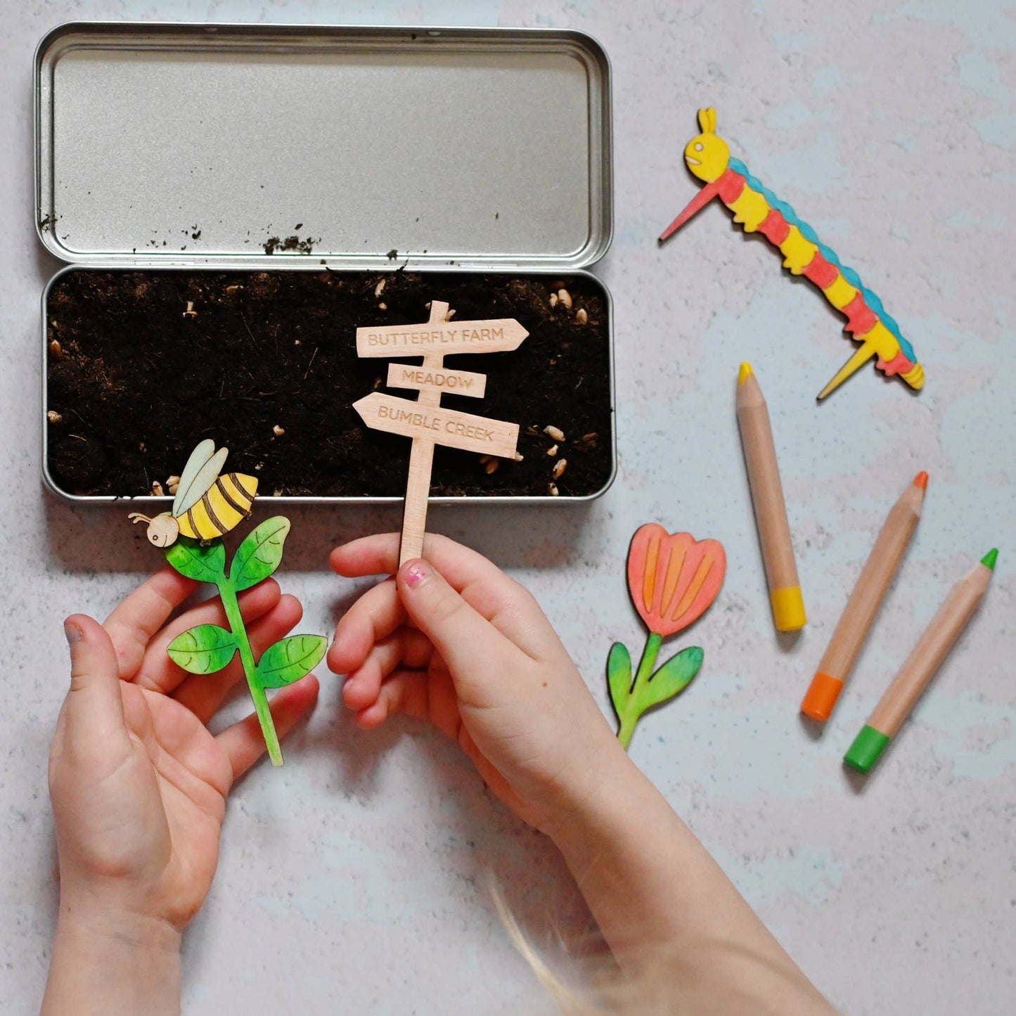 Children's hands holding a wooden stick with a small sign next to colorful markers and a toy dragonfly on a light surface.