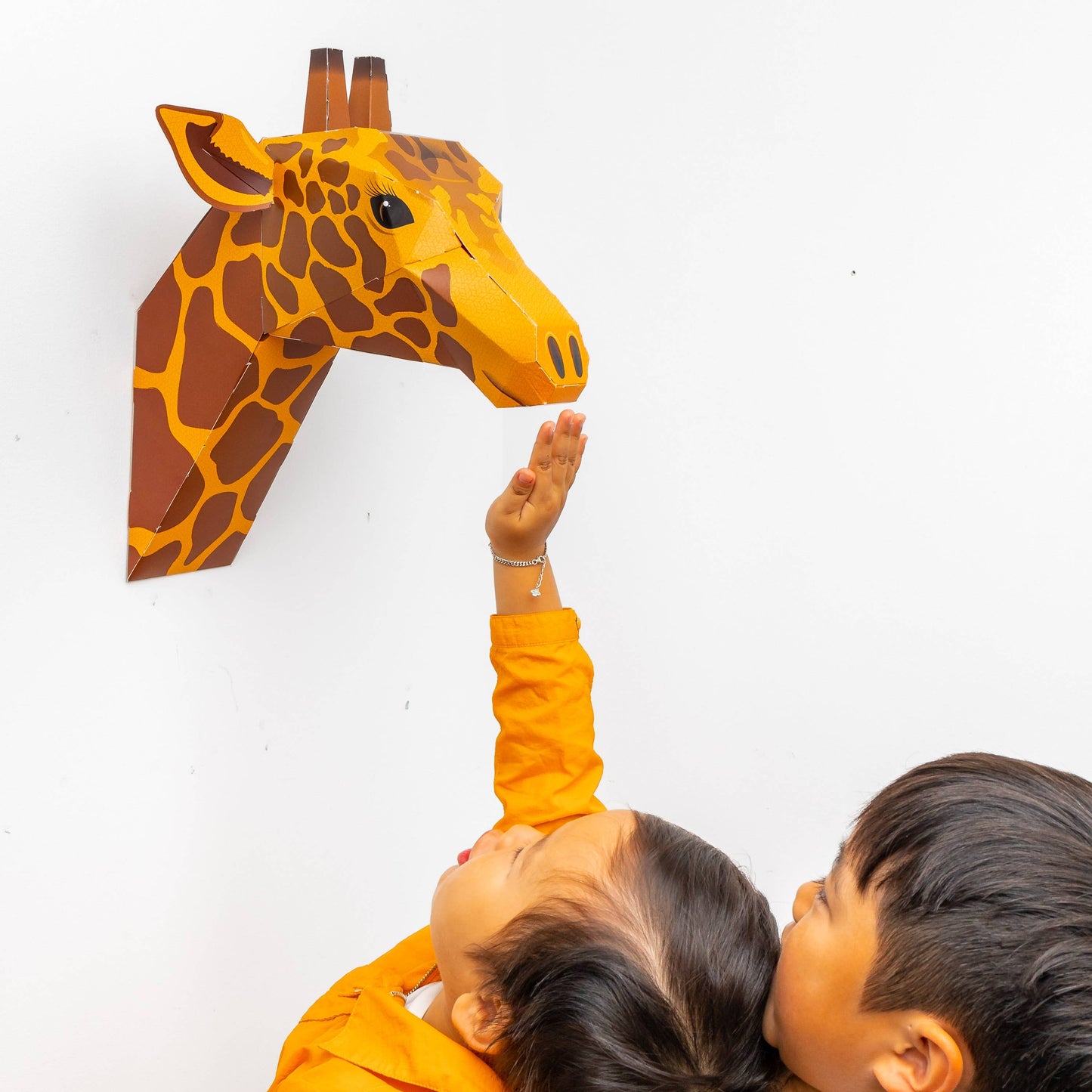 Two children reaching up to touch a giraffe-shaped head on a white wall.