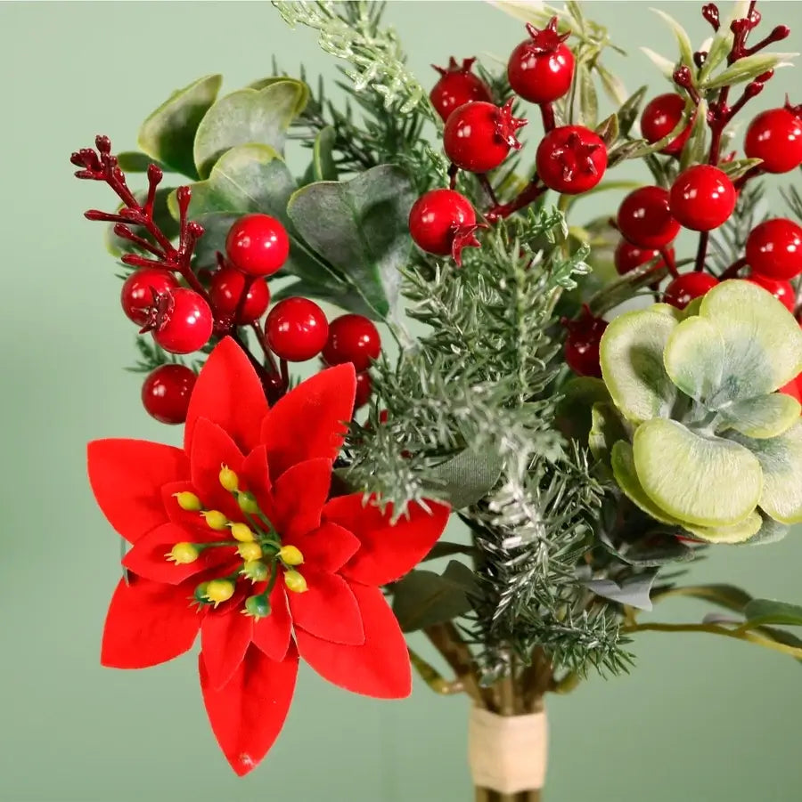 An artificial holiday floral arrangement featuring a bright red poinsettia, glossy red berries on red stems, silver-green pine branches, and pale green leaves, tied with a beige band at the bottom against a soft green background.