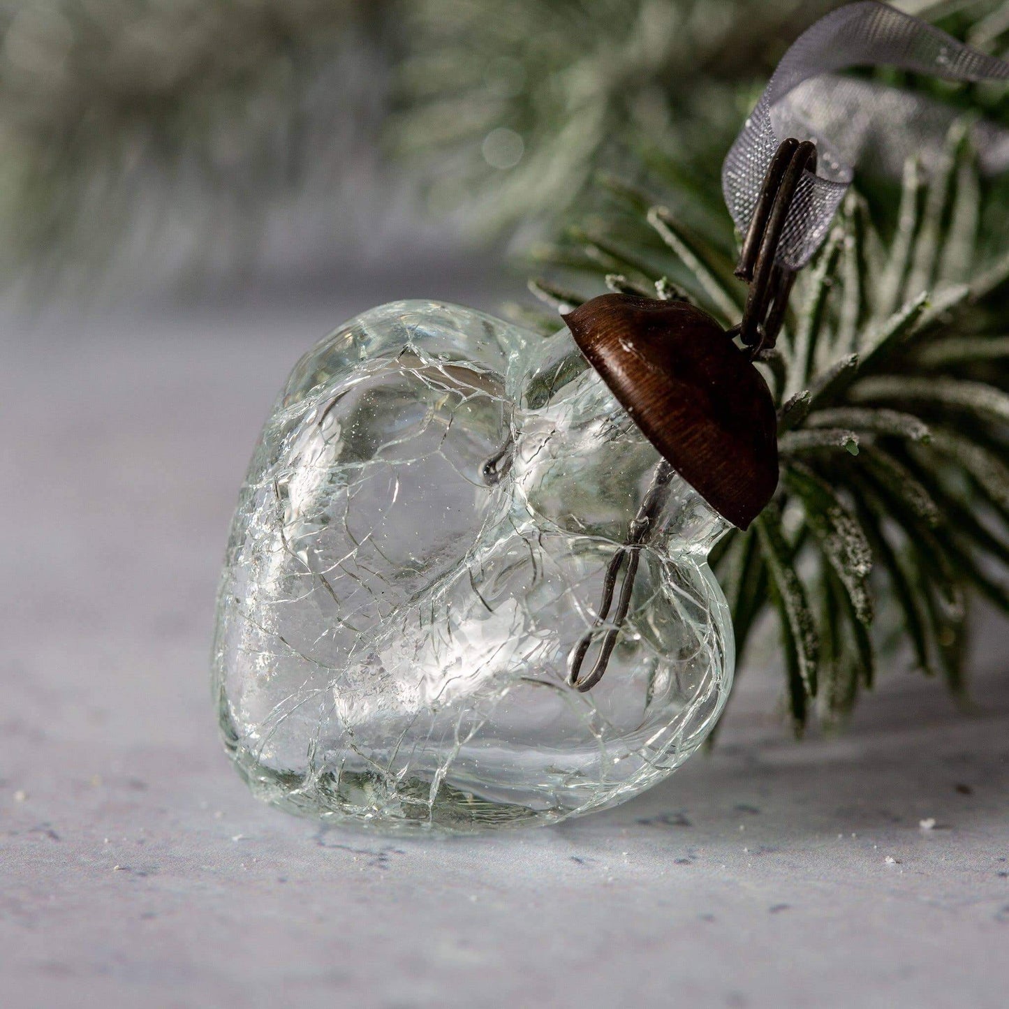 A clear crackle glass heart ornament with a bronze cap and organza tie, placed on a surface with a blurred greenery background.