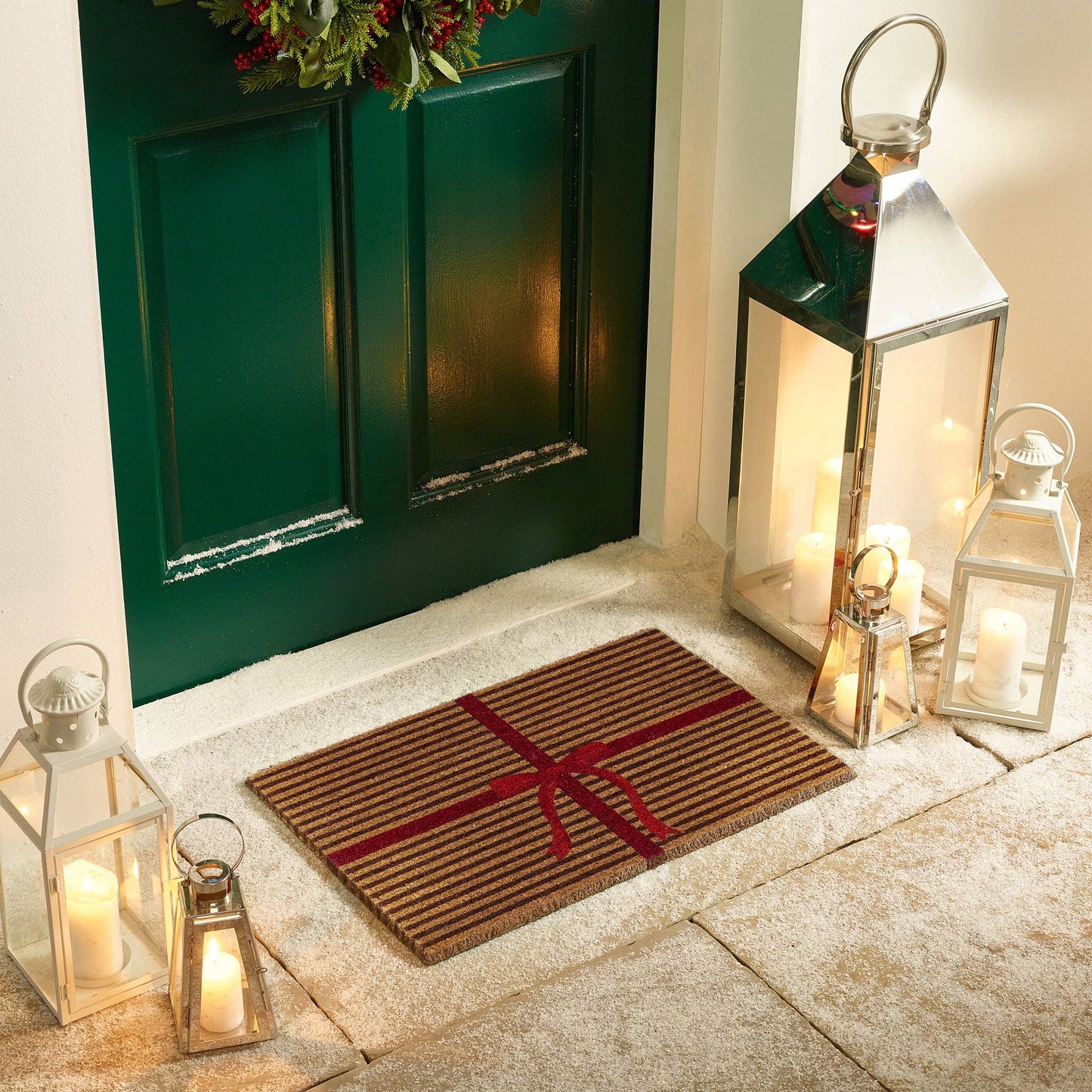 Decorative doormat with a red bow on a stone floor in front of a green door, surrounded by lanterns and candles.