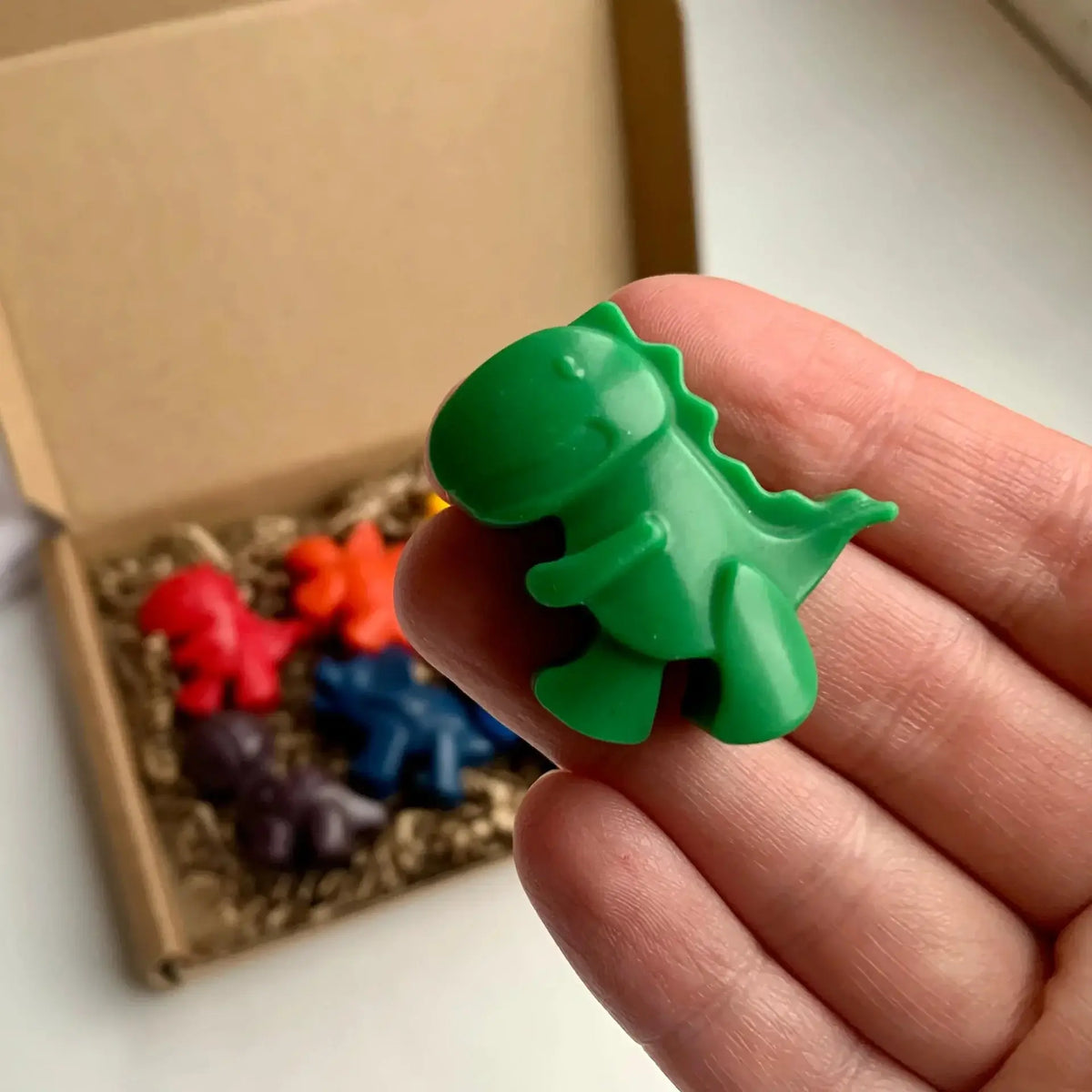 Green dinosaur-shaped crayon held by a hand with a box of crayons in the background.