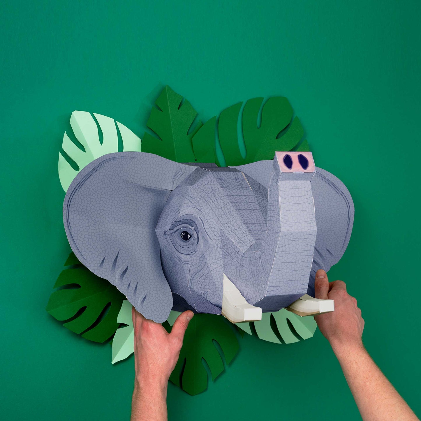 Gray elephant head decoration held by hands against a green background with leaf shapes