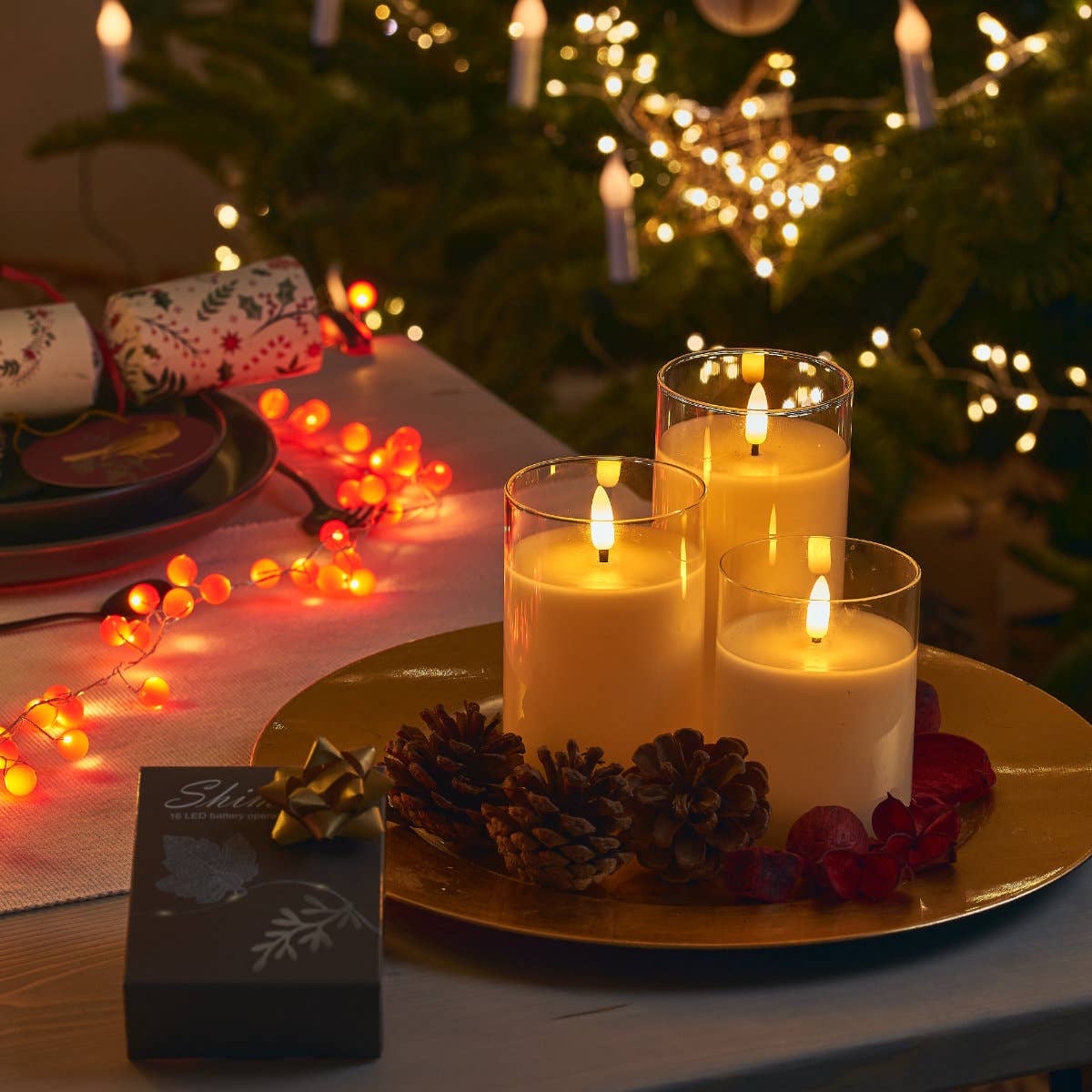 Decorative setup with candles, pinecones, and lights on a table in front of a Christmas tree.