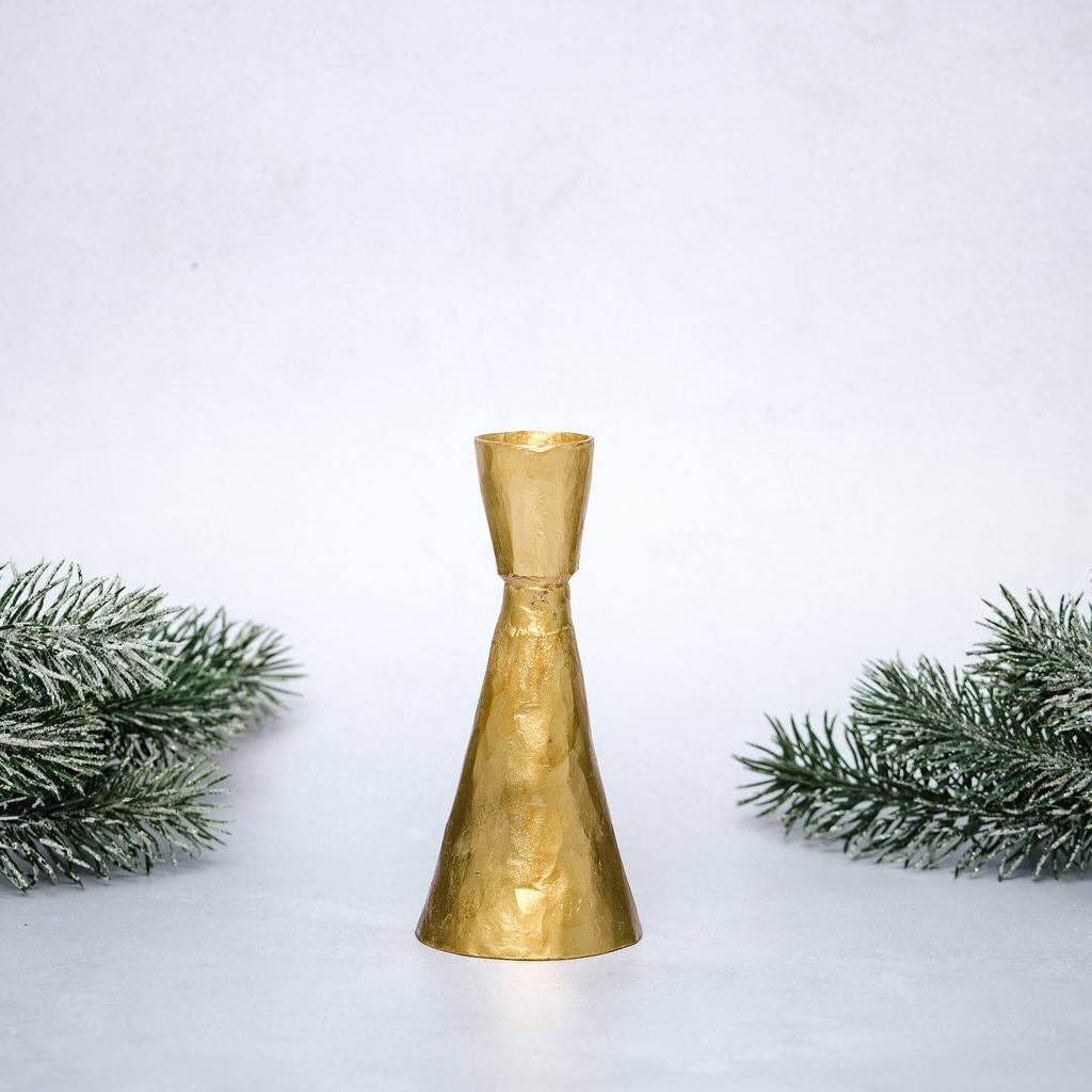 A gold colored metal candlestick holder of large size displayed on a white surface with green foliage at the sides.
