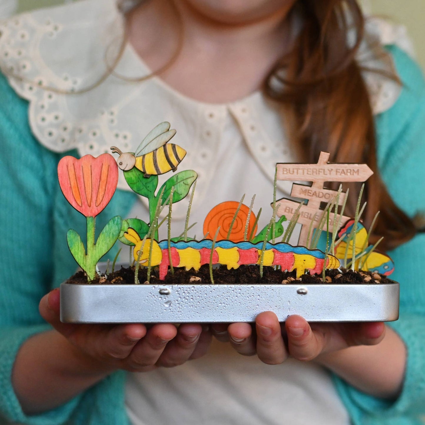 Child holding a small garden model with colorful elements and a bee illustration.