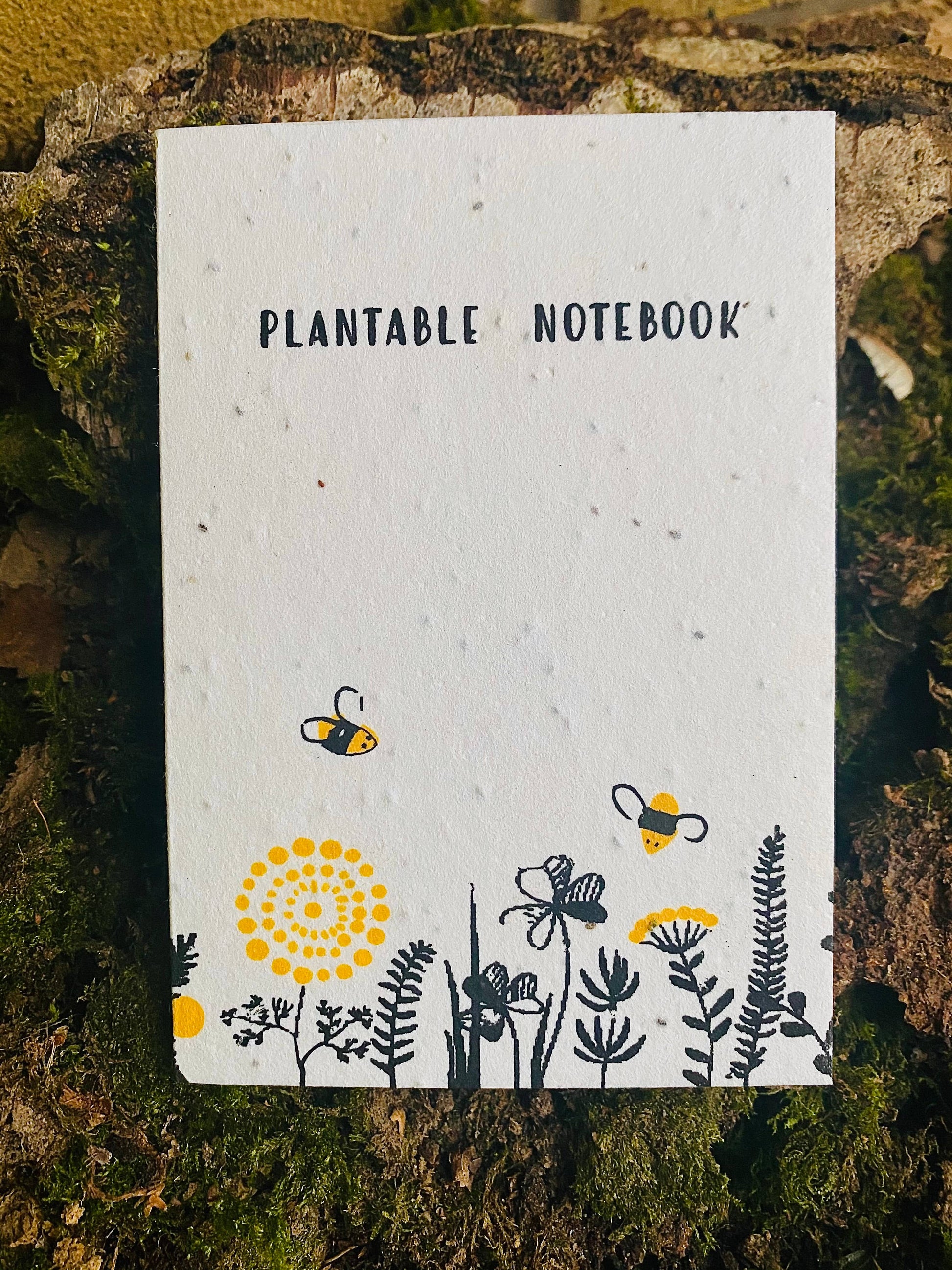 A white A6 notepad with a plantable design featuring black and yellow bee illustrations, labeled 'Plantable Notebook'.