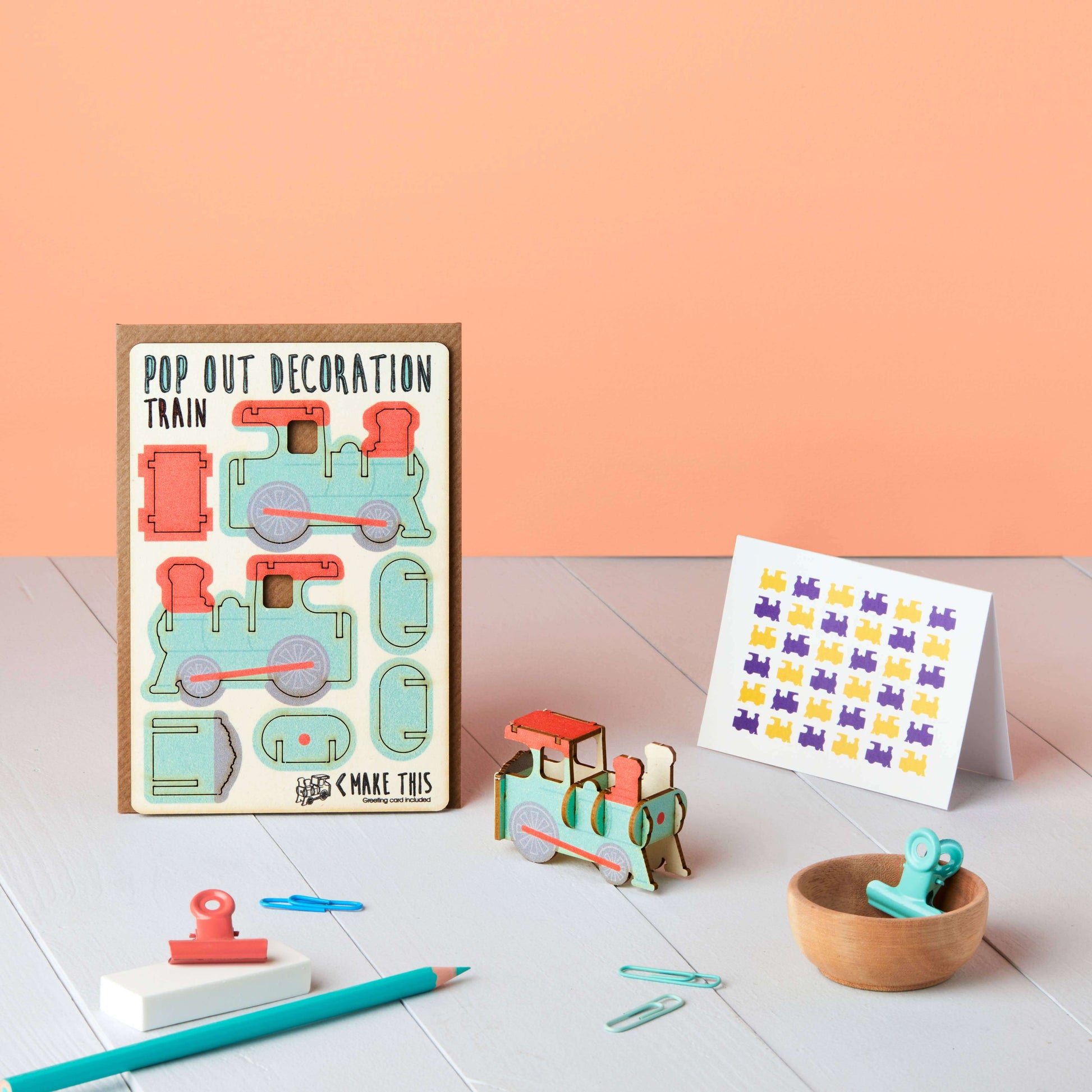 A greeting card with a 3D pop-out train design, accompanied by an A7 greeting card with a repeat train pattern and a brown A6 envelope. Both items have a cardboard train decoration with cut-out pieces sitting on a table