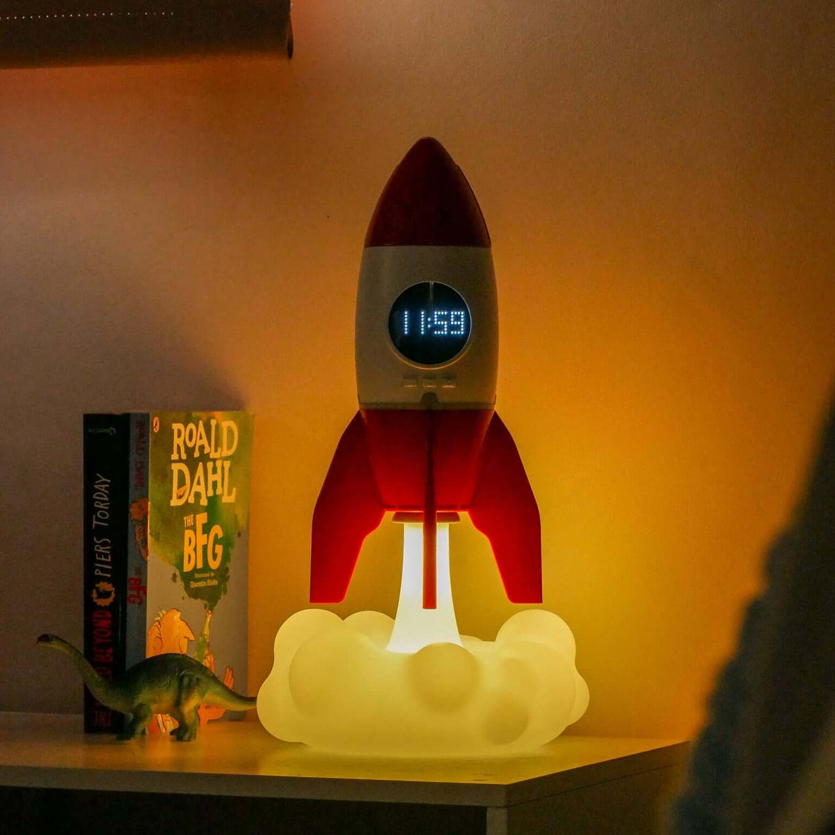 Rocket-shaped night light on a shelf with a book and toy dinosaur