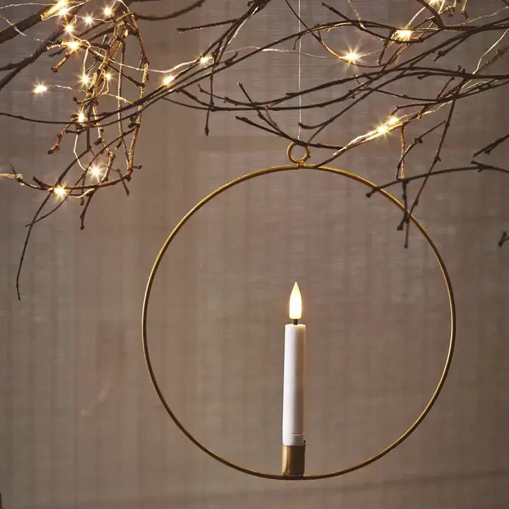 An LED candle ring with a rustic gold finish, hanging from a branch, with a lit candle inside it, surrounded by warm white LED lights.