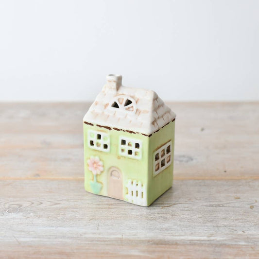 A small pastel green LED house with flower decorations and star cutouts on the roof, set against a wooden backdrop.