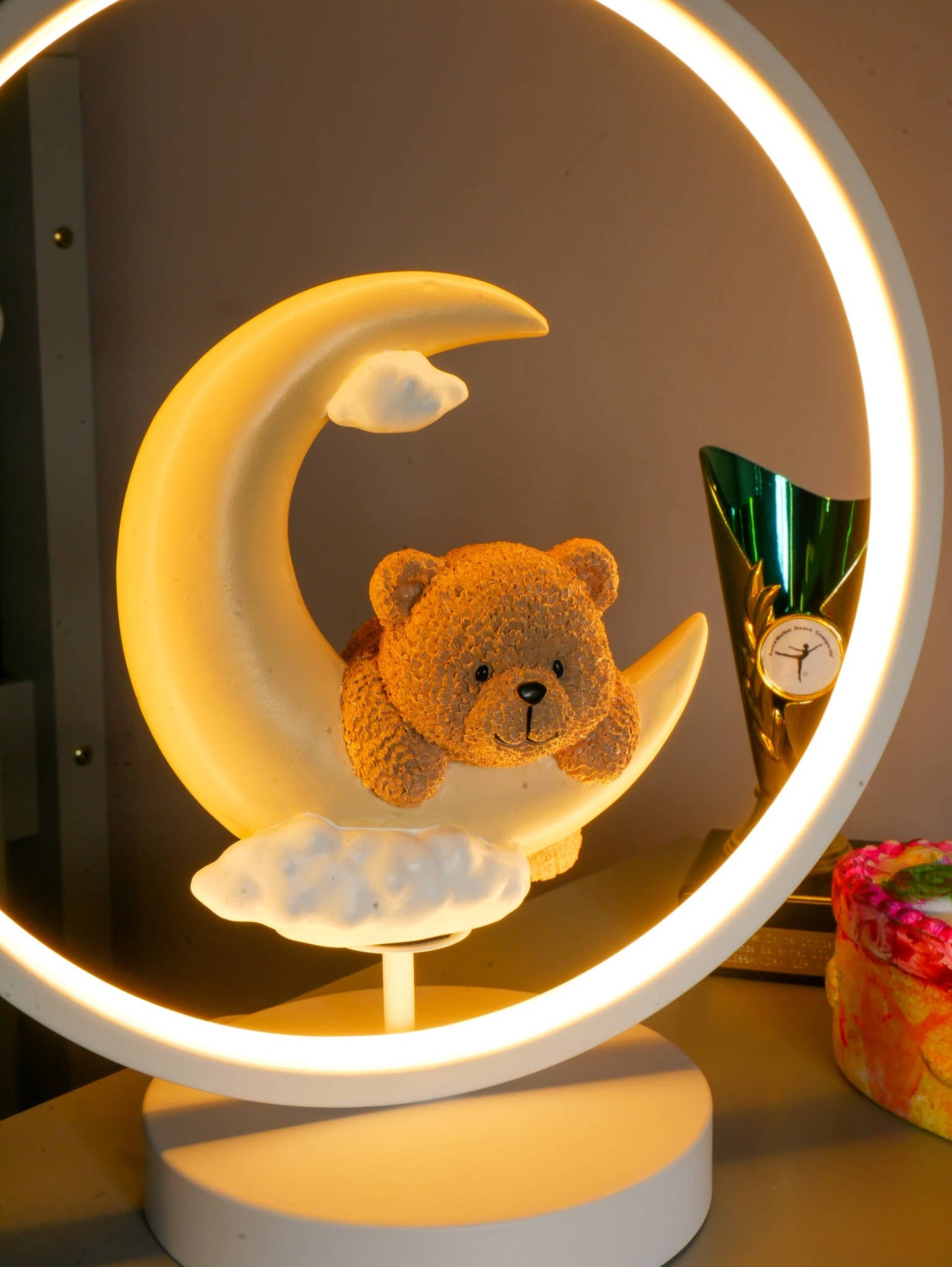 Teddy bear on a crescent moon with a circular light around it on a shelf.