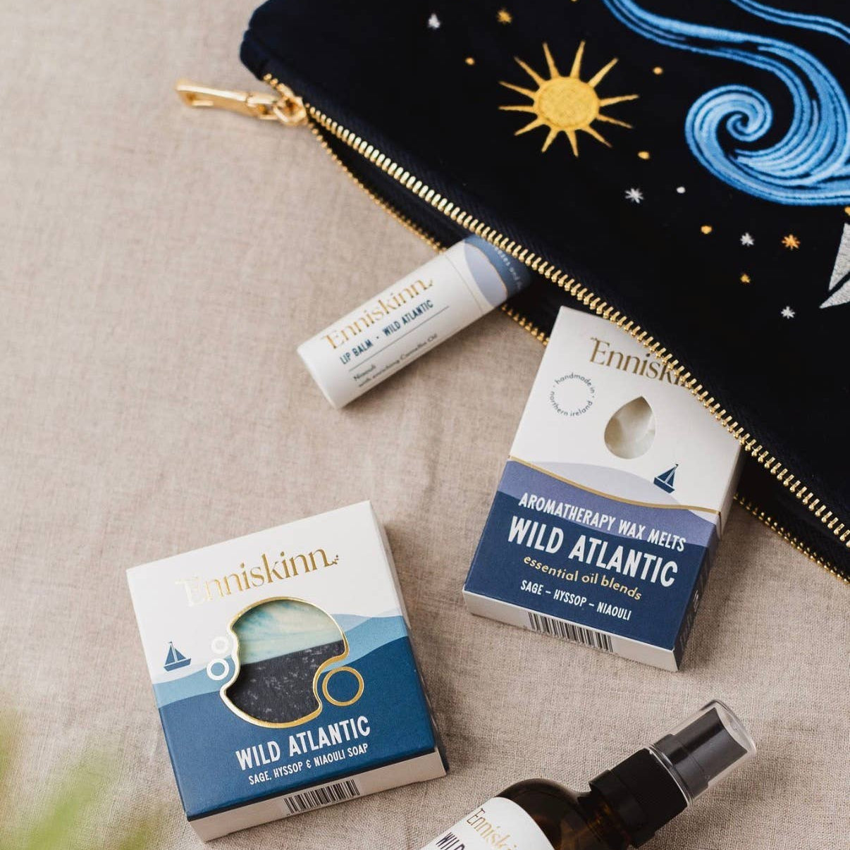 A flat-lay arrangement of Enniskinn 'Wild Atlantic' products on a beige linen background, including a navy blue zip pouch with embroidered stars and waves, a lip balm tube, two boxed soap bars, and a small spray bottle.