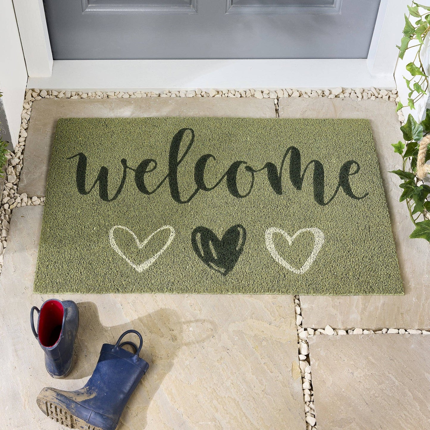Green doormat with 'welcome' and heart designs on a stone porch.