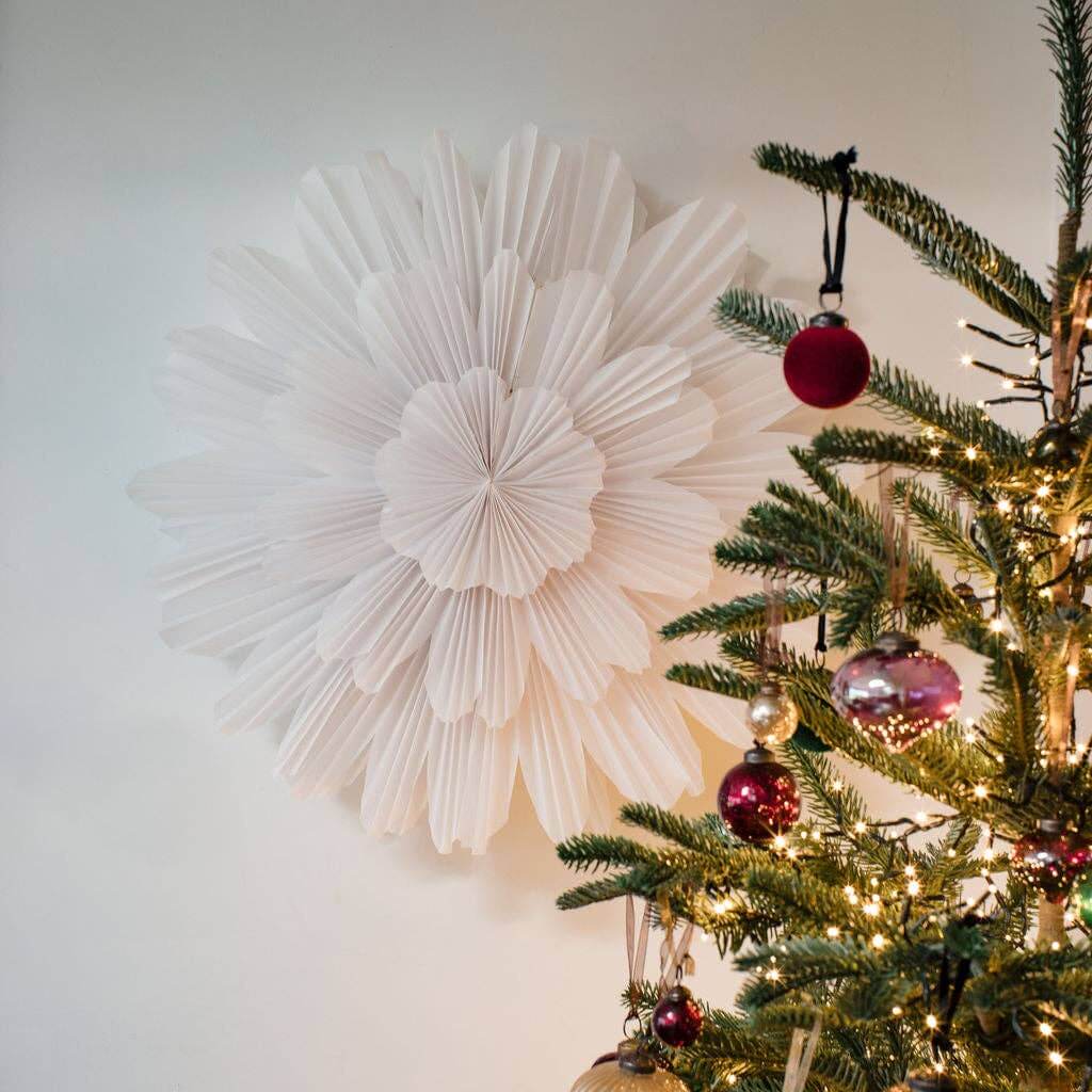 A large white paper wall hanging decoration with a geometric pattern, displayed above a Christmas tree with a natural green branch.