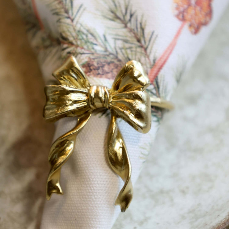 A close-up of a shiny gold bow-shaped ring holding a white napkin with a holiday pine branch and orange flower print, resting on a textured light surface.