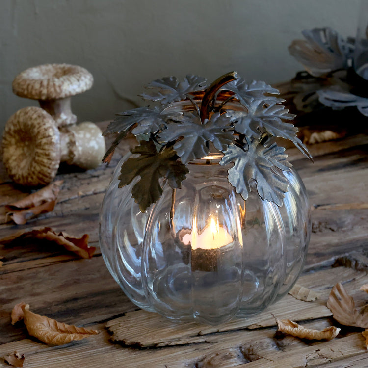 A clear ribbed glass pumpkin-shaped candle holder with a lit tea light inside, decorated with faux silver-gray leaves and a brown stem, set on a rustic wooden surface with dried leaves and mushrooms in the background.