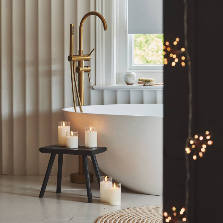 Flameless LED pillar candles sitting on a small wooden table beside a modern white curved bath in a minimalist bathroom