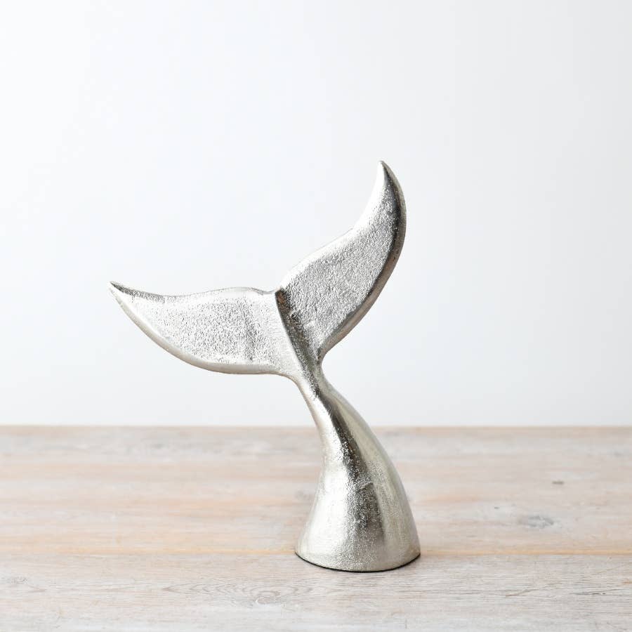 Metallic silver whale tail sculpture on a light wooden surface with a white background