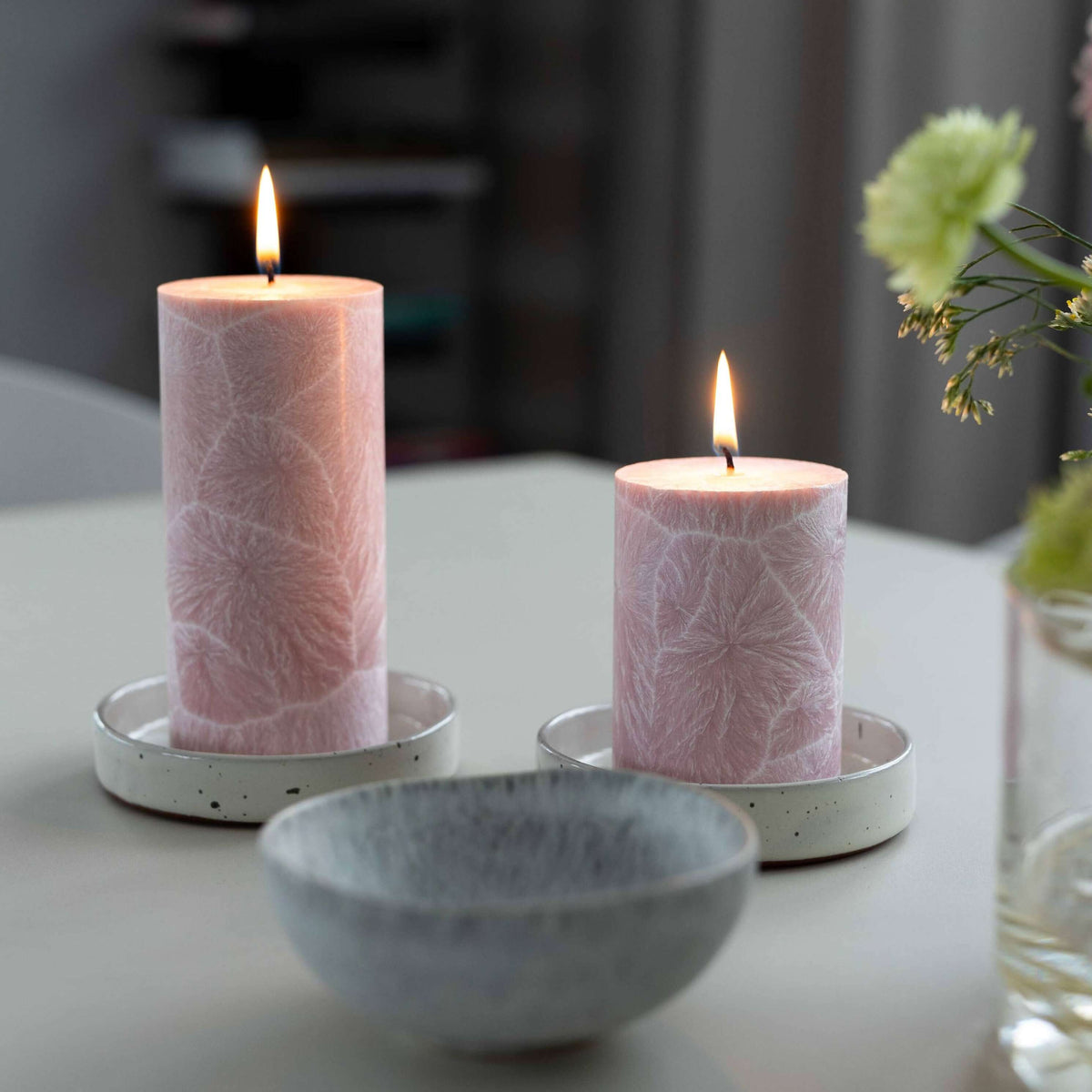Two lit pink floral pillar candles in white ceramic holders on a light gray table, with a small gray bowl in the foreground and blurred flowers on the right.