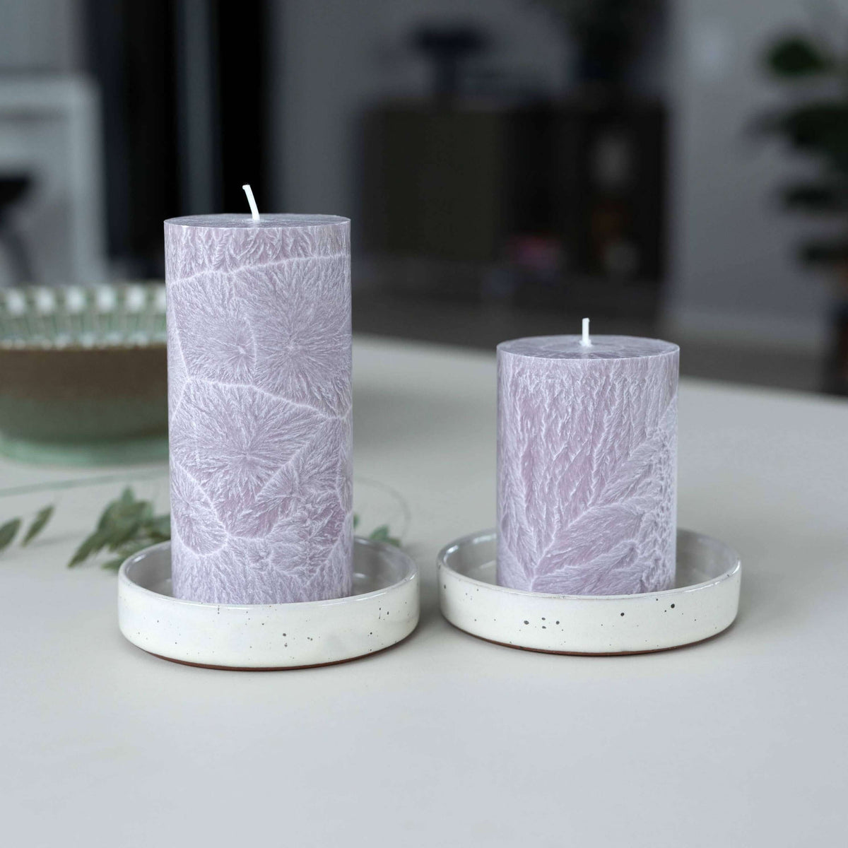 Two lavender pillar candles with a leaf pattern, displayed in white speckled ceramic holders on a light tabletop.