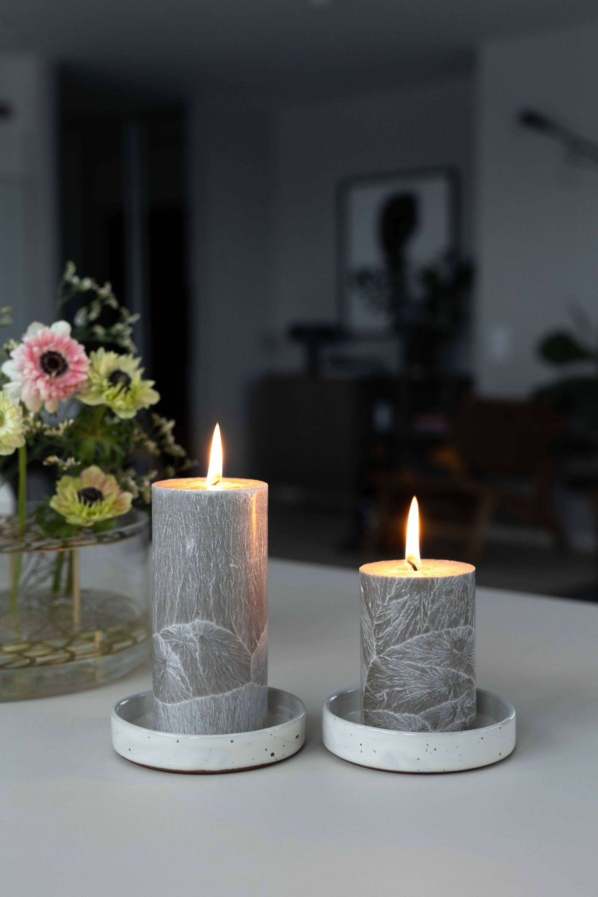 Two lit pillar candles in round holders with a textured gray and white marble-like pattern, displayed on a tabletop in a softly blurred indoor setting.