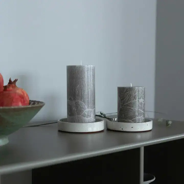 Two gray pillar candles with a white leaf pattern resting on white ceramic saucers on a dark shelf, with a bowl of pomegranates partially visible on the left.