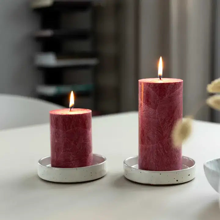 Two lit pillar candles in a matte white ceramic holder, set on a light gray tabletop with a softly blurred indoor background.
