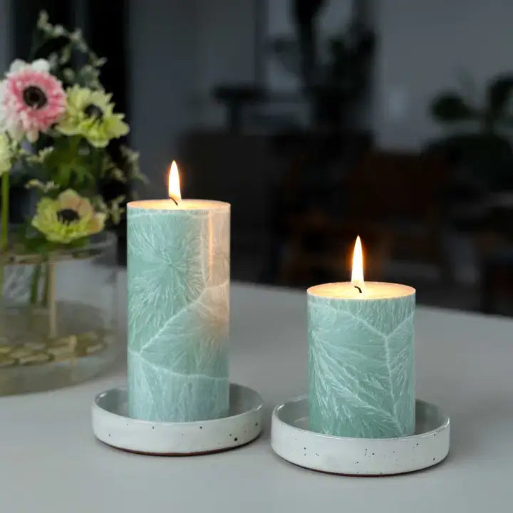 Two lit pillar candles with a pale mint green and white leaf pattern, sitting in matching speckled white ceramic holders on a tabletop. A blurred vase of flowers is visible in the background.