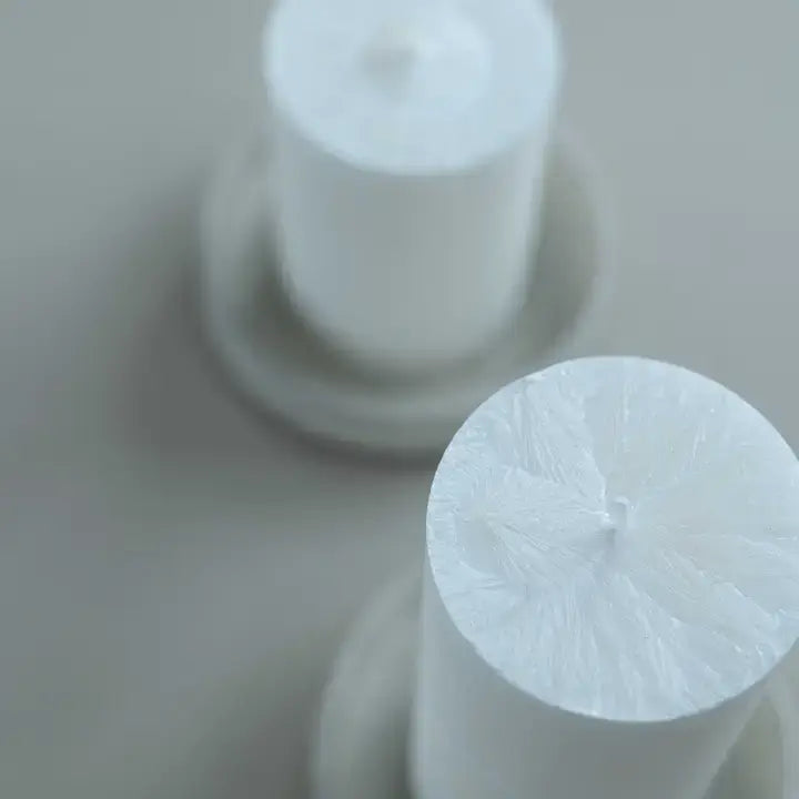 Two white pillar candles in matching white holders, with one candle in the foreground sharply in focus and the other softly blurred in the background.