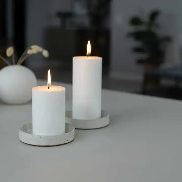 Two white pillar candles with flames, resting on matching round white holders on a light gray tabletop, with a softly blurred indoor background.