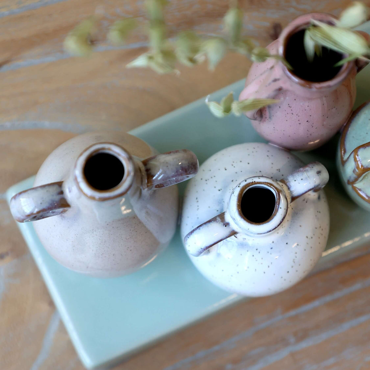 Two small ceramic vases on a light green square plate with a shell and additional vases in the background.
