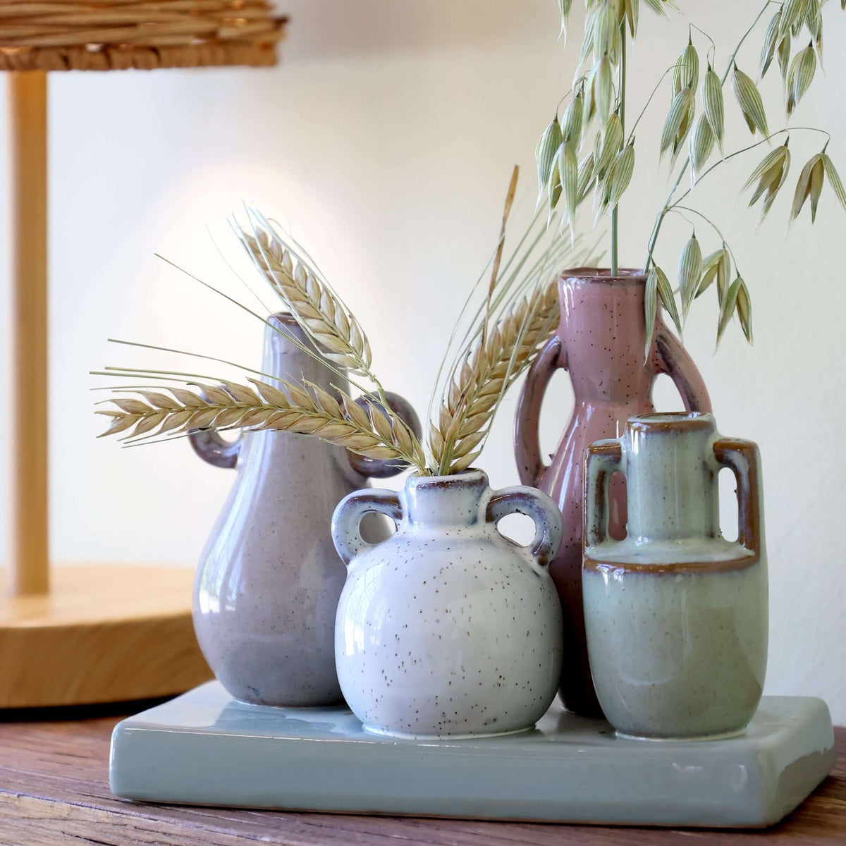 Set of ceramic vases on a wooden surface with a lamp and plant in the background
