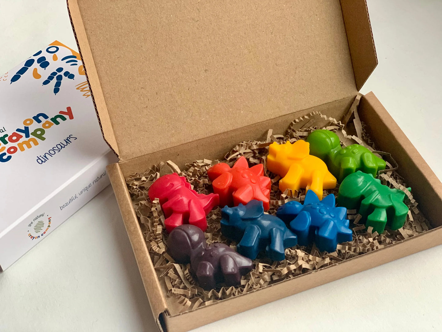 Colorful dinosaur-shaped crayons in a cardboard box with a brand box in the background.