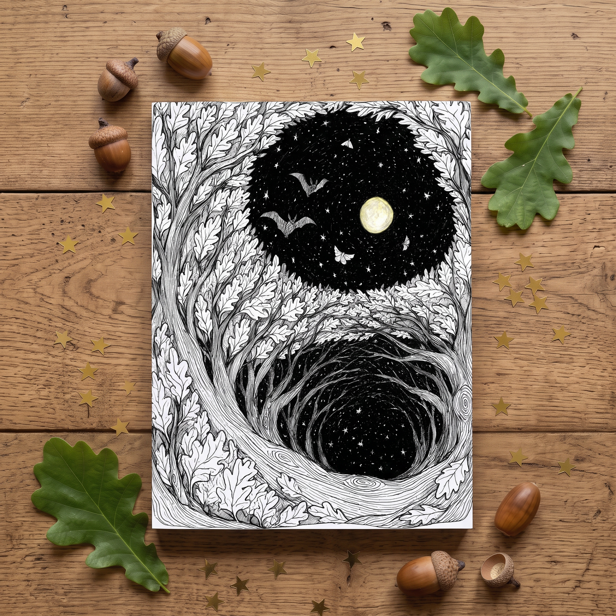 A notepad featuring a black and white line art illustration of two forest moon scenes, styled flat-lay on a wooden surface with oak leaves, acorns, and gold star confetti.