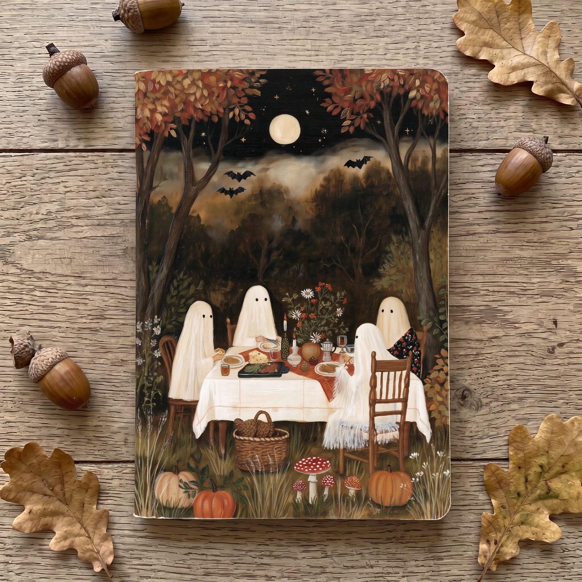 A notepad featuring a Halloween woodland scene with three ghost figures seated at a dinner table, surrounded by pumpkins, mushrooms, bats, and autumn trees under a full moon. The print is photographed from above on a rustic wood surface, with acorns and leaves placed around it.