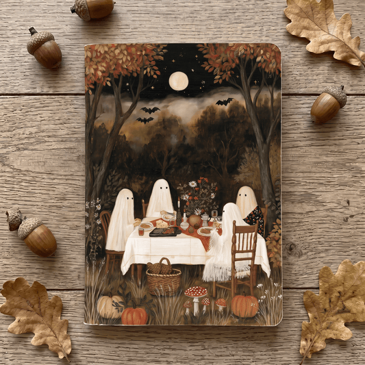 A notepad featuring a Halloween woodland scene with three ghost figures seated at a dinner table, surrounded by pumpkins, mushrooms, bats, and autumn trees under a full moon. The print is photographed from above on a rustic wood surface, with acorns and leaves placed around it.