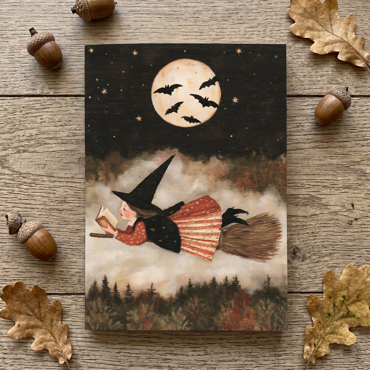 A notepad featuring a witch riding a broomstick under a full moon with bats, styled on a rustic wood surface with scattered acorns and leaves around it.