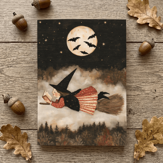 A notepad featuring a witch riding a broomstick under a full moon with bats, styled on a rustic wood surface with scattered acorns and leaves around it.