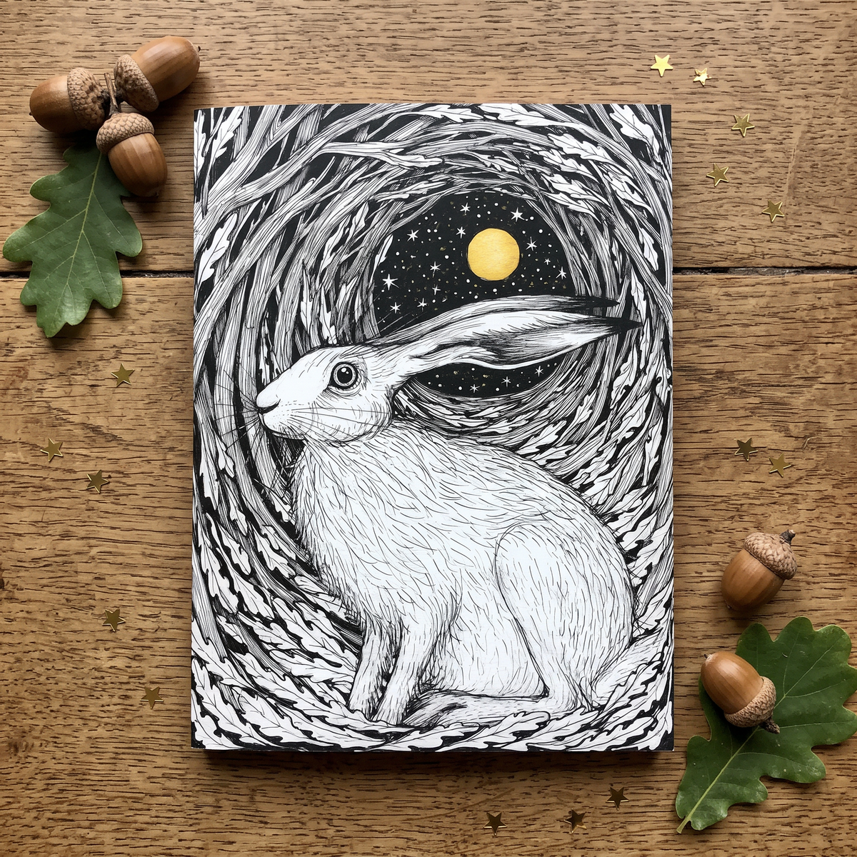 A notepad cover featuring a detailed black and white illustration of a hare sitting in a circular nest under a starry sky with a yellow moon, photographed on a wooden surface with acorns and leaves around it.