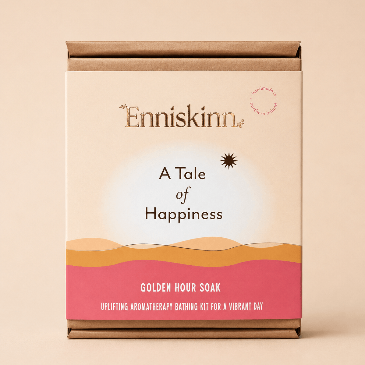 A boxed bath kit labeled 'Enniskinn' and 'A Tale of Happiness' with the variant name 'Golden Hour Soak,' shown against a warm beige background. The package is kraft brown with a cream center panel, black text, and a pink lower band with wavy orange lines.