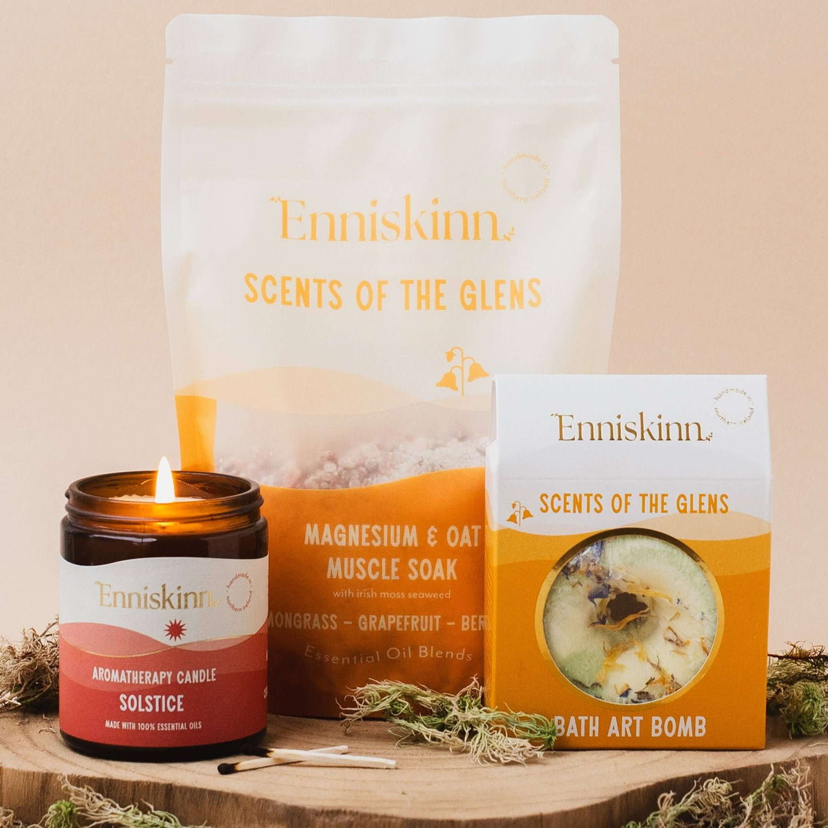 Enniskinn 'Scents of the Glens' bath and body gift set featuring a white and orange pouch of magnesium & oatmeal muscle soak, a red and amber atharotherapy candle, and a boxed bath art bomb, arranged on a wooden slice with dried moss against a beige background.