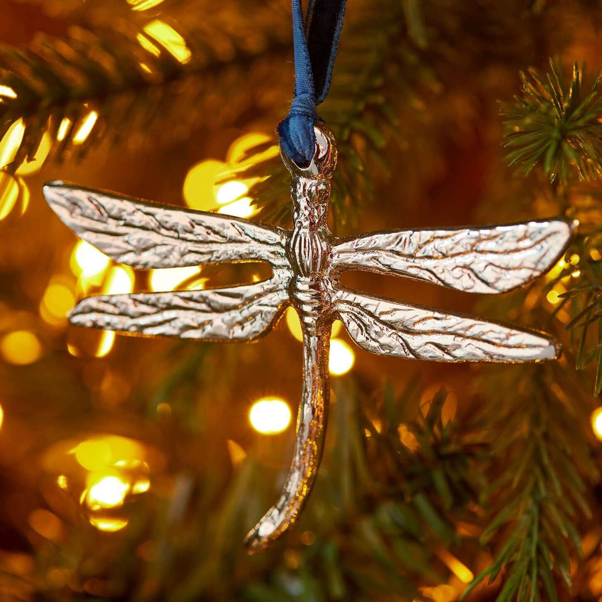A metallic dragonfly Christmas ornament with a textured silver body and wings, hanging from a dark blue ribbon on a decorated Christmas tree with warm lights.